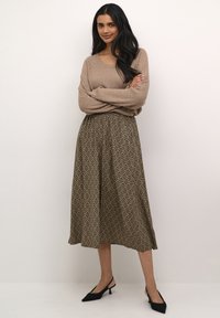 Beige bordás pulóver, amelyet egy barna mintás midi szoknyával párosítanak, amely geometrikus mintázattal rendelkezik. A modellen fekete slingback magassarkú cipők vannak.