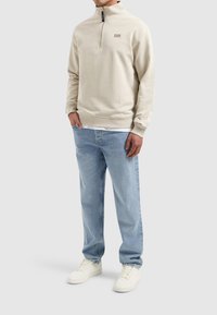 Lichtbeige sweatshirt met een halve rits kraag en logo-detail, gecombineerd met lichtblauwe jeans en witte sneakers. Casual fit.