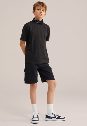 Fiú áll, a háta mögött összekulcsolt kézzel, fekete pólót, fekete zsebes rövidnadrágot, fehér zoknit, fekete-fehér tornacipőt és fekete nyakkendőt visel.