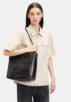 Schwarze Tasche aus Krokoleder mit breitem Boden, Schulterriemen und minimalem Branding an der Unterseite. Glatte Textur, strukturierte Form.