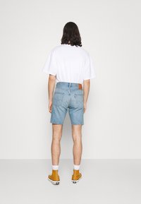 Hombre con el cabello a la altura de los hombros que lleva una camiseta blanca, pantalones cortos de mezclilla azul claro, calcetines blancos y zapatillas altas de color mostaza.