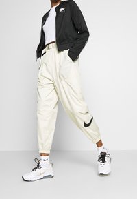 Osoba v bílých teniskách Nike, krémových cargo kalhotách s černým swooshem, bílých ponožkách, bílém zkráceném topu a černé zipové bundě Nike.