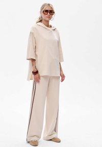 Beige hoodie met een zachte textuur en een col, gecombineerd met beige wijde broek met donkere strepen aan de zijkanten.
