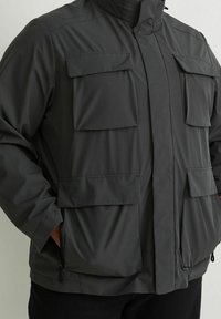 Giacca impermeabile grigia con colletto alto, diverse tasche frontali e chiusure con zip, realizzata in tessuto liscio con texture opaca.