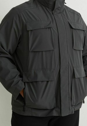 Grijze waterdichte jas met een hoge kraag, meerdere voorzakken en ritssluitingen, gemaakt van gladde stof met een matte textuur.