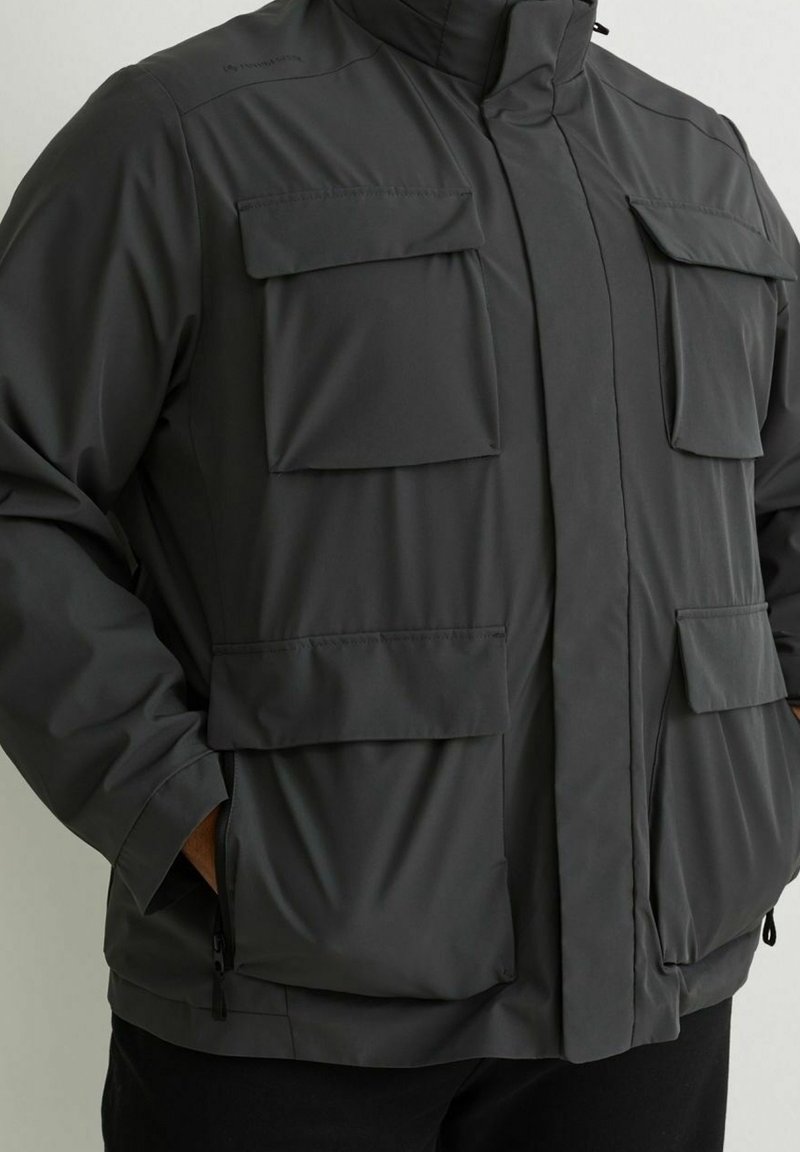 Giacca impermeabile grigia con colletto alto, diverse tasche frontali e chiusure con zip, realizzata in tessuto liscio con texture opaca.