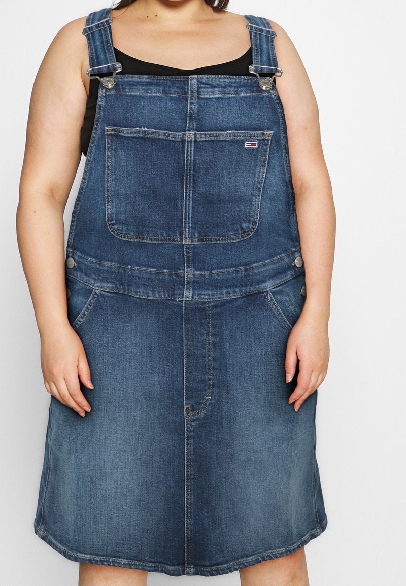 Denimová šaty na trakaře ve středně modré barvě, s nastavovacími ramínky, přední kapsou a kapsami po obou stranách.