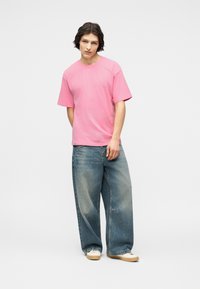 Νεαρό άτομο με σκούρα μαλλιά μήκους μέχρι τους ώμους, φοράει φωτεινό ροζ κοντομάνικο πουκάμισο, φαρδιά μπλε τζιν και λευκά αθλητικά, στέκεται μπροστά σε απλό φόντο.