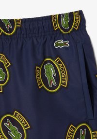 Bañadores de baño en color azul marino con un diseño de logo de Lacoste repetido, cinturilla elástica y un bolsillo lateral. Fabricados con un tejido ligero y de secado rápido.