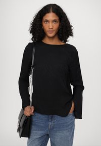 Svart langermet skjorte med teksturert mønster, med rund hals og sideslisser, sammen med høyhalsede blå jeans og en svart veske.