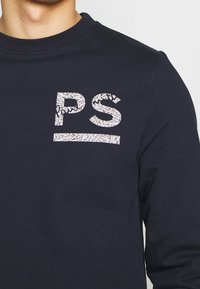 Sudadera azul marino con cuello redondo circular, que presenta un logo "PS" blanco texturizado y una franja horizontal. Fabricada con un tejido suave y con un acabado liso.