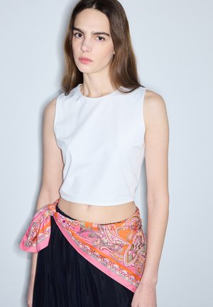 Jonge vrouw draagt een wit mouwloos crop top, een zwarte rok en een roze paisley sjaal gestrikt rond haar middel tegen een effen achtergrond.