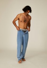 Helle blaue Denim-Jeans mit einem lässigen Schnitt, fünftaschen Design und Knopfverschluss, getragen mit einem schwarzen Markenbund und barfuß.