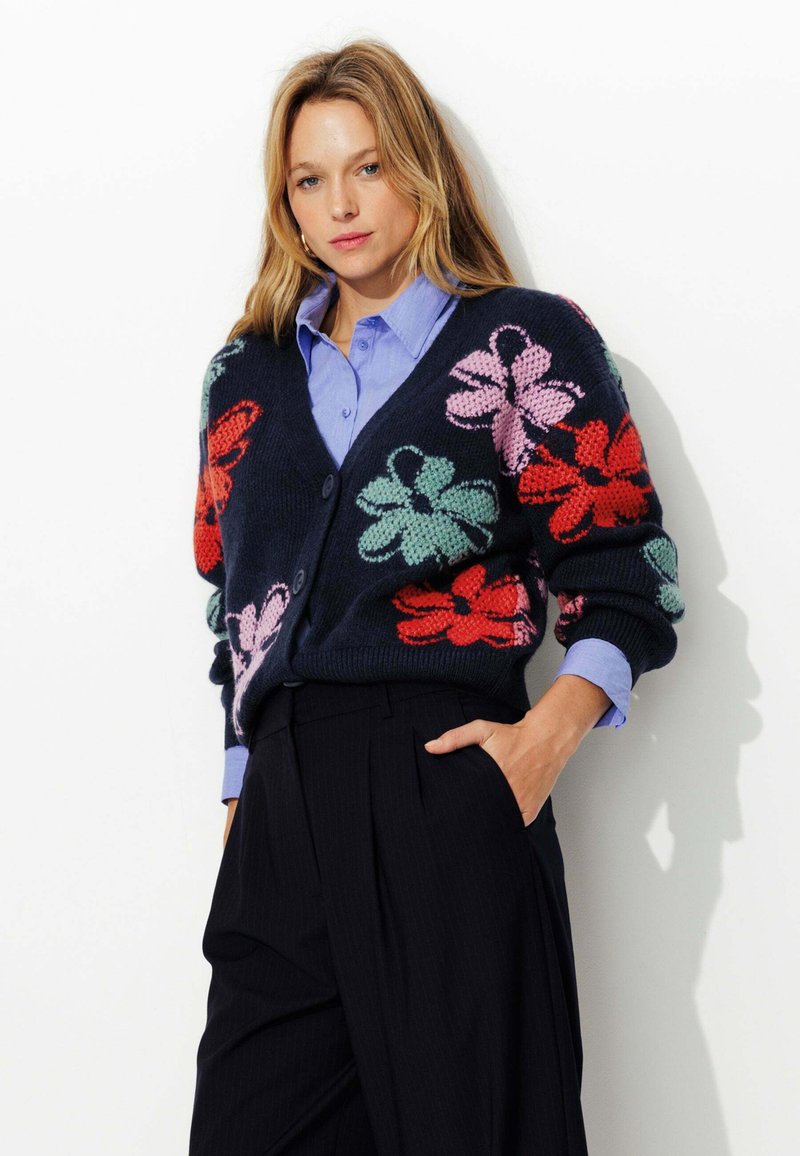Cardigan tricot marine avec de vives fleurs rouges, roses et vertes ; doté d'un col en V, de boutons et de poignets côtelés. Porté par-dessus une chemise bleu clair.