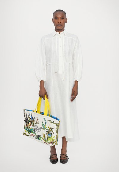 Abito bianco in lino con lunghe maniche a sbuffo e scollo con coulisse. Tiene una borsa colorata e decorata con manici gialli e dettagli blu.