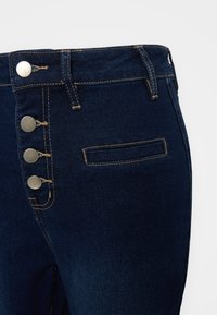 Pantalones de mezclilla azul oscuro con cuatro botones metálicos, costuras beige en contraste, pasadores para cinturón y un bolsillo recto horizontal en el lado derecho frontal.