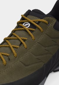 Αθλητικό παπούτσι σε ελιά πράσινο με μαύρη σόλα, κίτρινες και μαύρες κορδόνια με σχέδιο, μαύρο επενδυμένο πέλμα και λευκό κυκλικό λογότυπο στη γλώσσα.