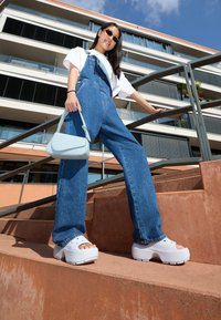 Zoccoli bianchi con punta rotonda e suola texture, salopette in denim con gambe wide e una borsa azzurra, sullo sfondo di un edificio moderno.