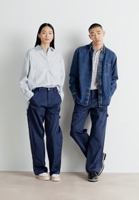 Casual outfit met een lichtblauw overhemd met knopen, marineblauwe cargobroek en een spijkerjack over een geruit hemd. Getextureerde stoffen zichtbaar.