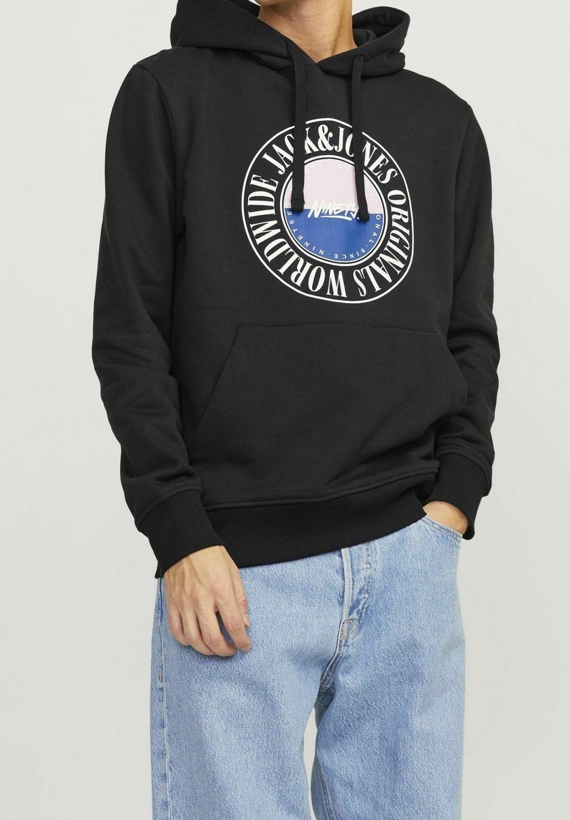Personne portant un sweat à capuche noir avec le logo Jack & Jones Originals Worldwide et un jean bleu clair, une main dans la poche et l'autre détendue.