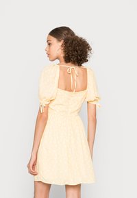 Vestido amarelo floral com mangas bufantes, costas abertas e laço na cintura. Feito de um tecido leve com um padrão branco subtil.