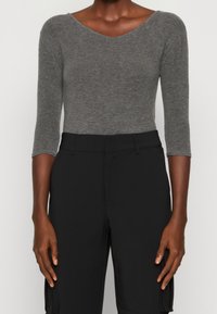 En person klädd i en åtsittande grå topp med trekvartsärmar som är instoppad i högmidjade svarta byxor, står med armarna avslappnat vid sidorna.