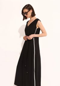 Czarna sukienka bez rękawów z dekoltem w kształcie litery V, z białą falbaniastą lamówką wzdłuż boku oraz regulacją w talii na sznurku.