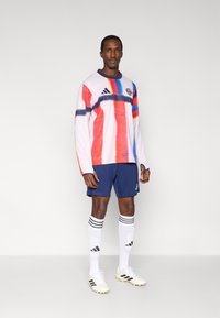 Ilgomis rankovėmis sportinis marškinėlis baltas su raudonu ir mėlynu abstrakčiu raštu, derinamas su tamsiai mėlynais šortais, baltomis kojinėmis su juodomis juostelėmis ir baltais futbolo bateliais.