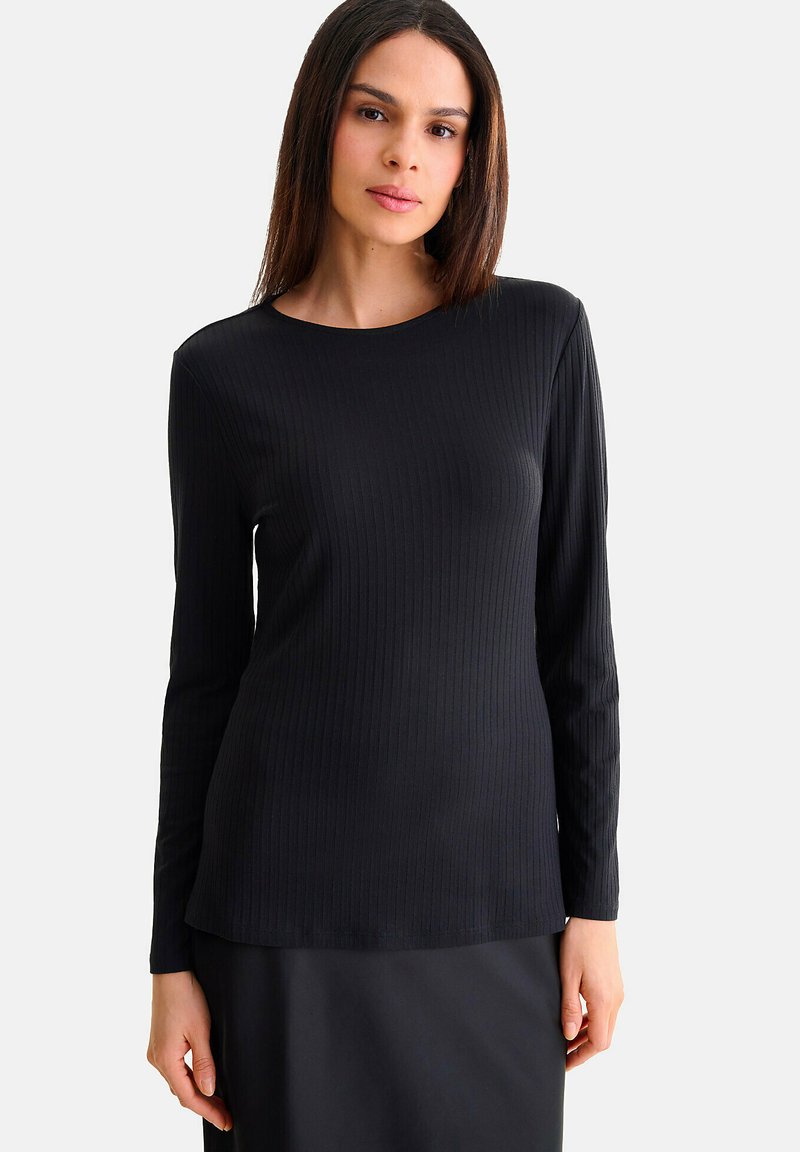 Haut noir à manches longues avec un col rond, texture lisse et ourlet légèrement arrondi, porté sur une jupe foncée assortie.