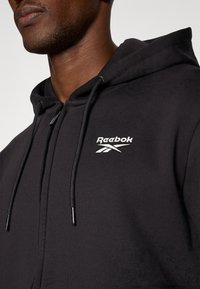 Črna Reebok kapuca z zadrgo, kapuco s vrvico in sprednjim logotipom. Gladka tkanina z oprijetim krojem in kovinskimi detajli.