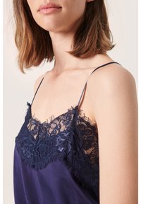 Camisola di satin blu navy con pizzo lungo il décolleté e le spalline, caratterizzata da una consistenza morbida e delicati dettagli frangiati ai bordi.