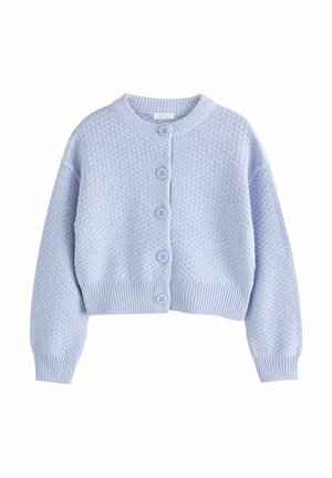 Enfant portant un cardigan boutonné texturé violet clair, un jean large en denim foncé avec de grands revers, et des baskets noires avec des bandes blanches, debout.