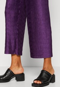 Personne portant un pantalon court texturé violet et des mules noires à talons blocs, avec les orteils et les chevilles visibles sur un fond uni.
