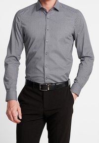 Hedvábná košile vzorovaná v černé a bílé barvě s límečkem na knoflíky, úzkým střihem, dlouhými rukávy a elegantním černým opaskem, doplněná o tmavé kalhoty.