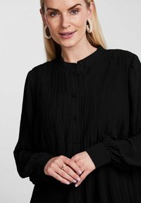 Blusa nera plissettata con scollatura rotonda, maniche lunghe e chiusura con bottoni. Presenta una texture liscia e una leggera lucentezza.