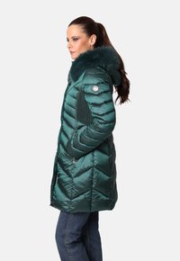 Mujer con coleta que lleva un abrigo acolchado verde oscuro con cuello de piel y detalles de punto en los lados, de pie de perfil con las manos en los bolsillos.