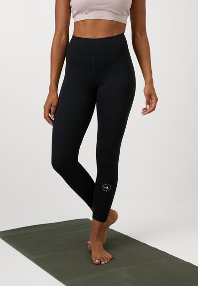 Magasan derekú fekete leggings, rugalmas anyagból készült, oldalsó varrásos kialakítással és kis emblémával az alsó lábszáron.