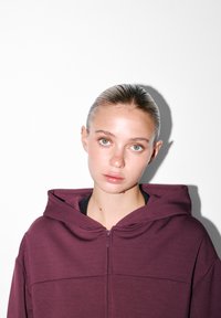 Mladá žena so slicked-back vlasmi, oblečená v zipsovom vínovom mikine, ktorá sa priamo pozerá dopredu na bielom pozadí so vrhnutým tieňom.
