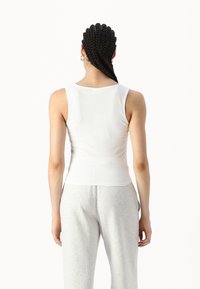 Hvit tettsittende tank topp med rund hals og uten ermer, laget av glatt stoff. Bært med grå joggebukser, som viser en enkel stil.