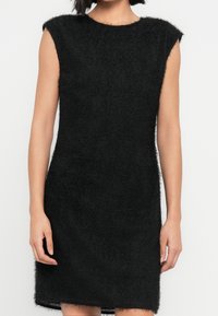 Robe sans manches en maille duveteuse noire avec col rond, portée par une personne visible des épaules jusqu'à mi-cuisse sur un fond uni.