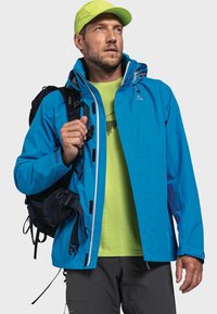 Blauwe waterdichte jas met een capuchon, witte ritssluiting en ventilatieroosters. Draagt over een neon groene shirt. Een zwarte rugzak is ook zichtbaar.