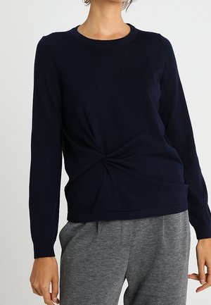 Frau trägt einen marineblauen Langarm-Pullover mit einer vorderen Wickeldetail und graue Freizeithose, steht vor einem schlichten Hintergrund.