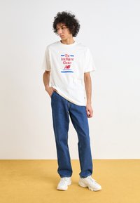T-shirt de algodão branco com texto impresso em vermelho e azul, combinado com calças de ganga azuis e ténis brancos, em pé sobre um fundo amarelo.