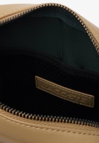Custodia con zip di colore beige, dalla finitura lucida, con un interno verde scuro e una tasca in rete nera, etichettata con un tag beige "LACOSTE".