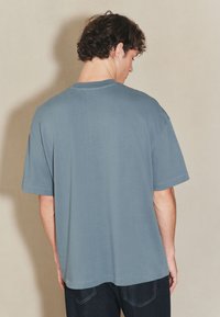 Tricou din bumbac gri, cu croială lejeră, mâneci scurte și guler rotund. Textură netedă și design simplu, fără ornamente.