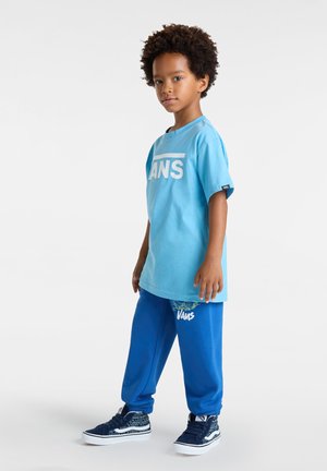 Hellblaues T-Shirt mit weißem "VANS"-Logo, kombiniert mit dunkelblauen Jogginghosen mit grafischem Druck und weißem "VANS"-Text. Dunkle Schuhe.