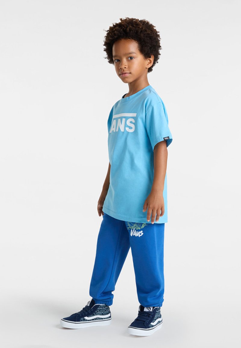 Lichtblauw T-shirt met wit "VANS" logo, gecombineerd met donkerblauwe joggingbroek met een grafische print en witte "VANS" tekst. Donkere schoenen.