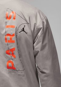 Grijze jasje met "PARIS" in vet oranje letters, een zwart Jordan-silhouetlogo en diverse teksten en grafische details op de achterkant.
