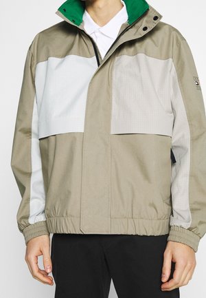 Mann trägt eine beige und hellgraue Jacke mit grün gefüttertem Kragen und weißem Poloshirt, steht mit entspannt an den Seiten hängenden Händen.