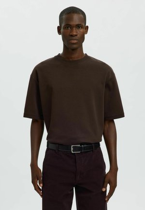 Jeune homme noir portant une chemise marron foncé oversize à manches courtes, une ceinture noire et un pantalon foncé, debout devant un fond clair uni.